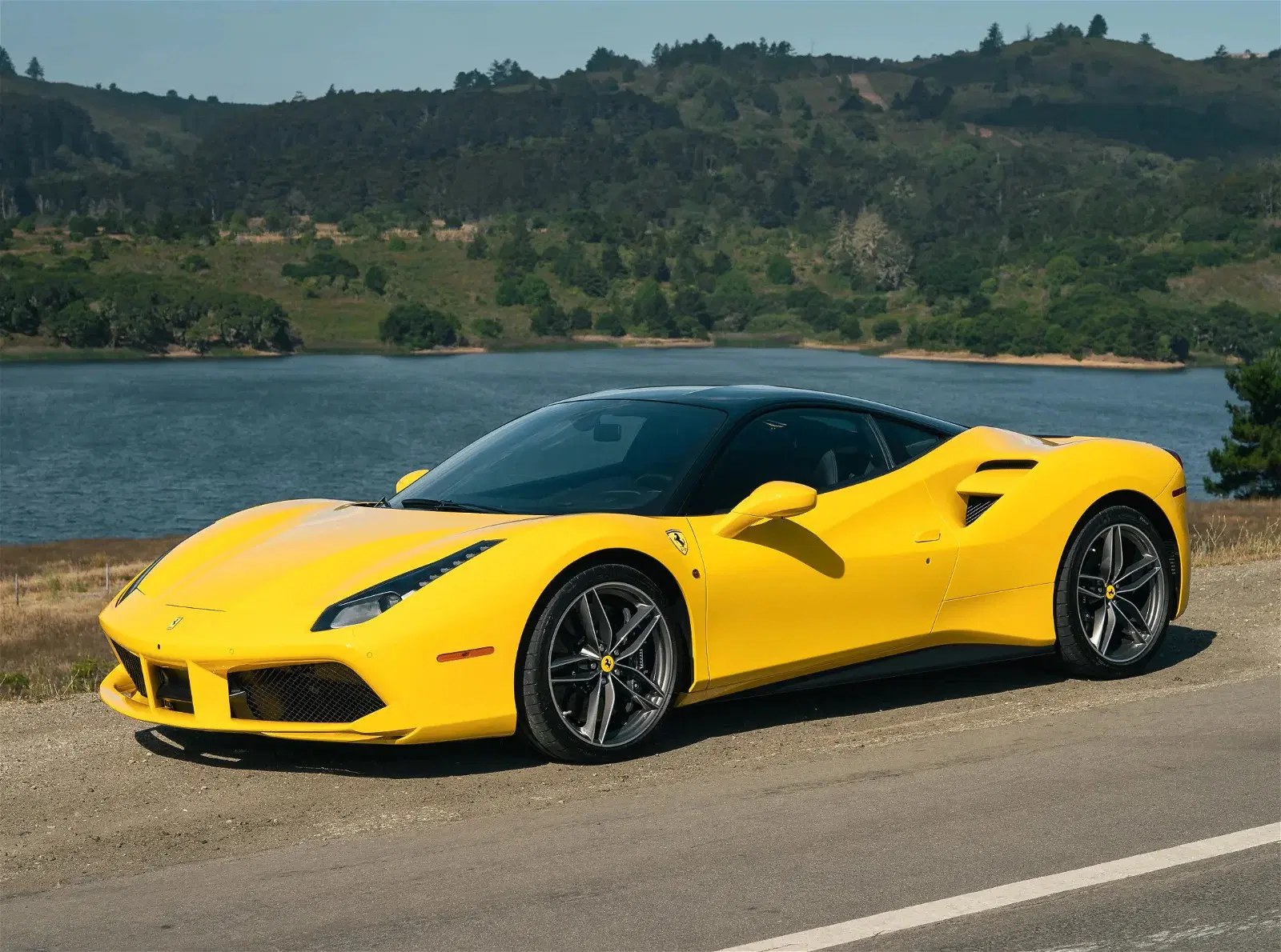 A yellow Ferrari parked by a lake with green hills behind